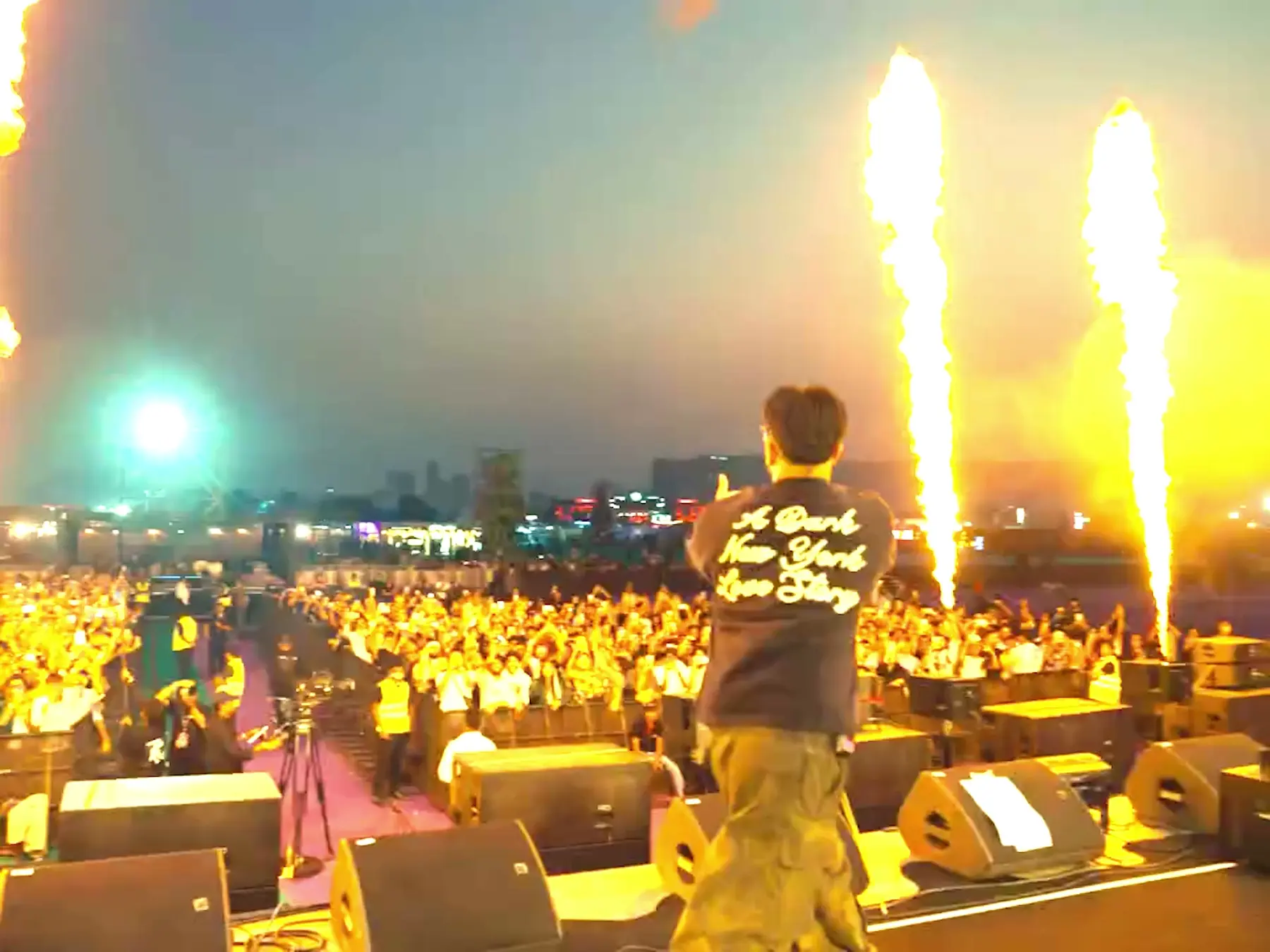 A rapper on stage between flame jets in front of a large crowd at dusk